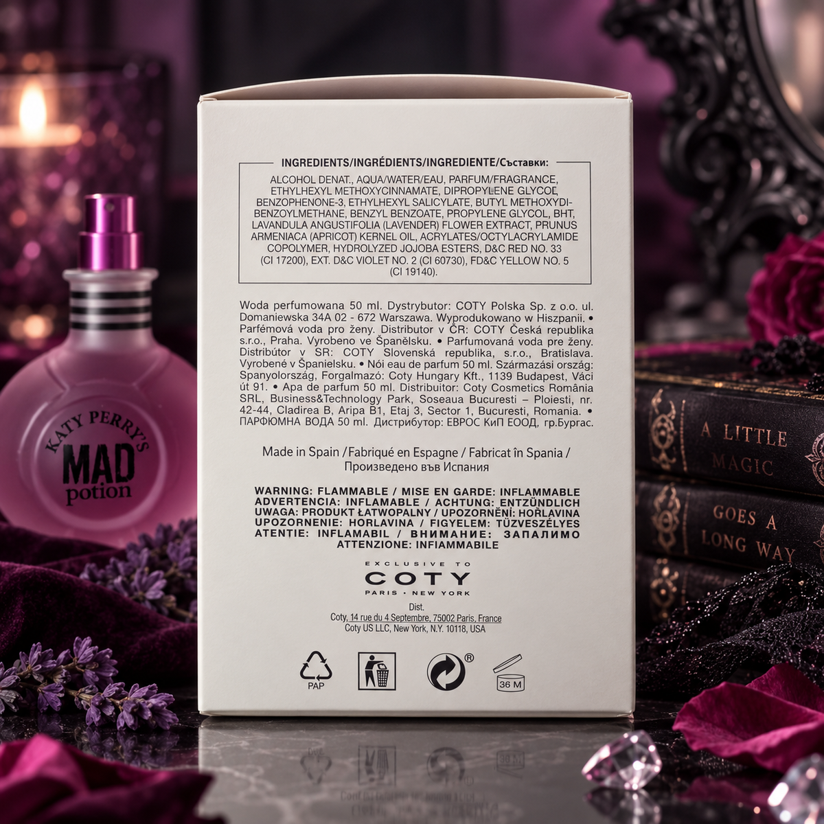 The back of a white Coty Paris perfume box is shown upright, with the brand name 'COTY PARIS' visible near the bottom. The box is photographed in a styled setting with purple flowers, a perfume bottle, and a decorative card in the foreground, against a softly blurred warm-toned background.